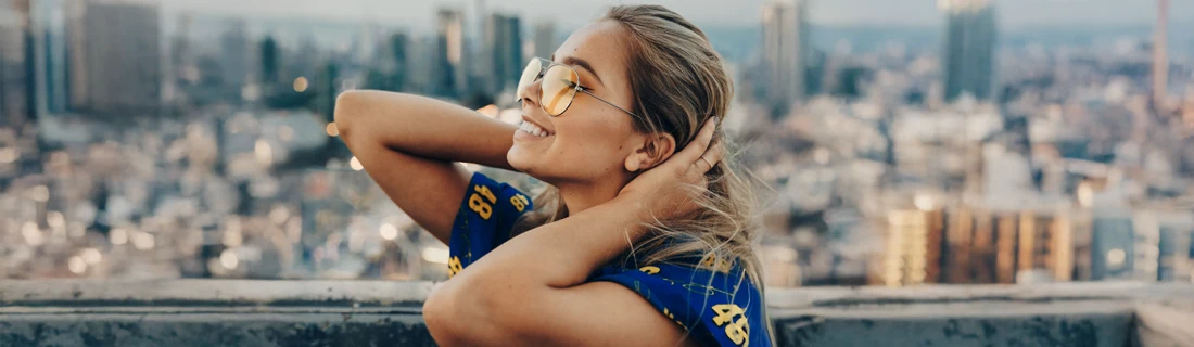 A woman in sunglasses, smiling with a city skyline in the background.